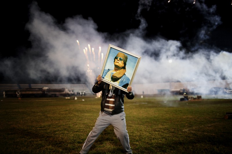 Libya, March 2011. A Qaddafi supporter holds a portrait of the Libyan leader as fireworks go up in the background on a soccer field in a suburb of Zawiyah where government minders took a group of foreign journalists to attend a staged celebration. Photo by Moises Saman/MAGNUM 